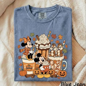May include: A blue t-shirt featuring a graphic of Minnie Mouse, Mickey Mouse, and Chip 'n Dale with pumpkin spice lattes and autumn leaves. The design includes text on the cups and a Comfort Colors label at the neckline. The shirt is short-sleeved.