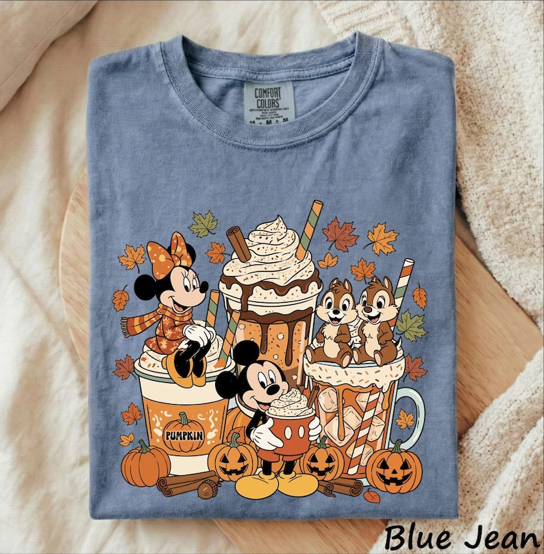 May include: A blue t-shirt featuring a graphic of Minnie Mouse, Mickey Mouse, and Chip 'n Dale with pumpkin spice lattes and autumn leaves. The design includes text on the cups and a Comfort Colors label at the neckline. The shirt is short-sleeved.
