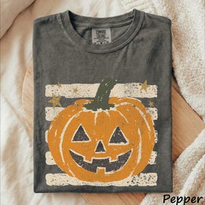 May include: A charcoal gray crew neck t-shirt featuring a graphic of an orange jack-o'-lantern pumpkin centered over horizontal white distressed stripes and gold stars. The inner neck label displays the text Comfort Colors in black lettering.
