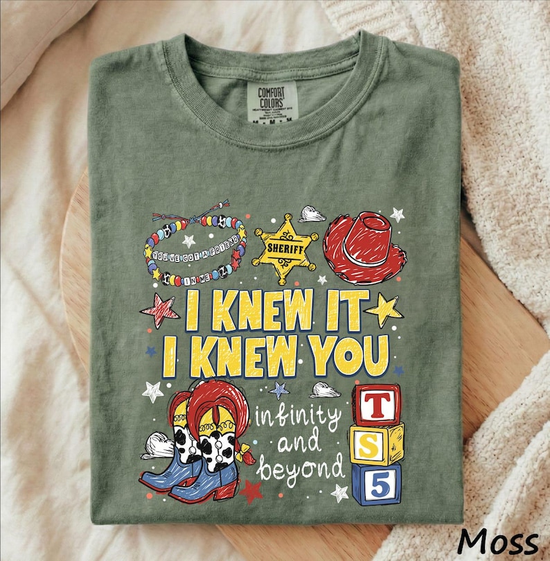 A green t-shirt with a colorful graphic design featuring cartoon characters, a cowboy hat, and various other elements. The text on the t-shirt reads "I KNEW IT I KNEW YOU" and "in infinity and beyond".