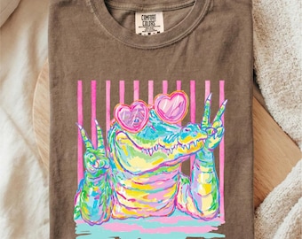 May include: Espresso-colored t-shirt featuring a colorful alligator wearing heart-shaped sunglasses and giving the peace sign. The graphic is set against a backdrop of pink vertical stripes. The shirt is made by Comfort Colors.