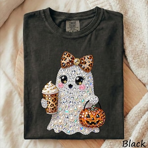 May include: A charcoal gray t-shirt featuring a graphic of a ghost covered in sparkling rhinestones. The ghost wears a leopard print bow, holds a leopard print coffee cup, and carries a glittery orange jack-o'-lantern bucket.