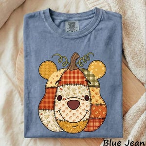 May include: A blue crew neck t-shirt with a Comfort Colors label at the inner neckline. The front features a graphic of a bear-shaped pumpkin made of various patchwork patterns in orange, yellow, and brown, including plaid and floral prints.