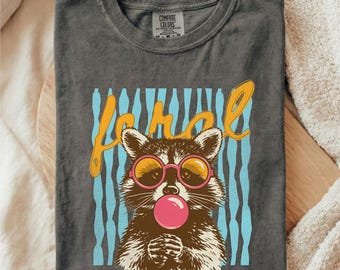 May include: Dark gray t-shirt with a graphic of a raccoon wearing pink sunglasses and blowing a bubblegum bubble. The word "Ferol" is written in yellow script above the raccoon, with blue vertical stripes in the background. The shirt is labeled "Comfort Colors".