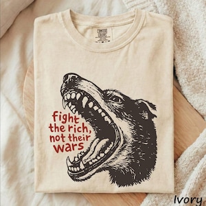 May include: An ivory crew neck t-shirt featuring a dark brown graphic of a snarling dog with its mouth wide open. To the left of the dog, the text reads in red: fight the rich, not their wars. The shirt has a Comfort Colors brand label at the neck.