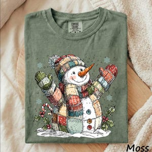 May include: A moss green t-shirt featuring a graphic of a snowman wearing a patchwork hat, scarf, and mittens. The snowman has an orange carrot nose, three buttons, and is surrounded by holly leaves and snowflakes. The label reads Comfort Colors.