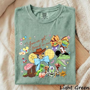 A green t-shirt with a colorful graphic design featuring various characters from the Toy Story franchise, including Woody, Buzz Lightyear, and other beloved characters.