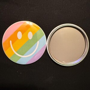 May include: A round, rainbow-colored compact mirror with a smiling face design. The mirror is open, revealing the reflective surface. The design features a rainbow of colors, including pink, orange, yellow, green, and blue.