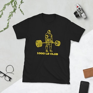 Może przedstawiać: Czarny T-shirt z żółtym konturem sztangisty podnoszącego sztangę. Pod obrazkiem nadrukowany jest tekst "1000 LB CLUB".