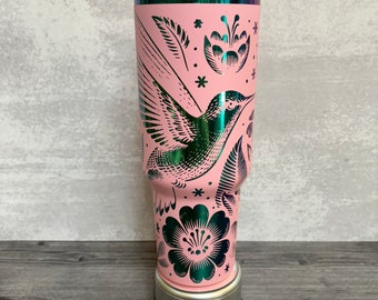 May include: Pink stainless steel tumbler with a hummingbird and floral design. The tumbler has a pink lid and a silver base.