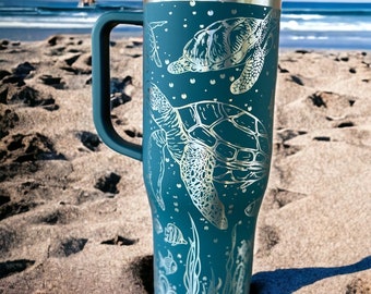 May include: A teal blue stainless steel travel mug with a handle. The mug features a design of sea turtles and seaweed in a silver colour.