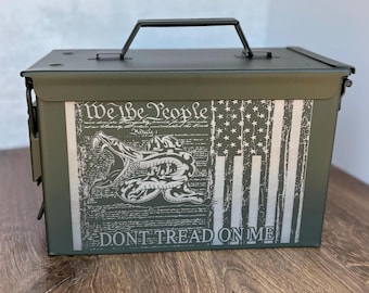 May include: An olive green metal ammunition box featuring a printed graphic on the front. The design includes the text We The People, a coiled rattlesnake, a stylized American flag, and the text DONT TREAD ON ME in a bold, sans-serif font.