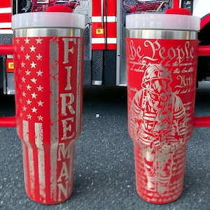 May include: Two red stainless steel travel mugs with lids and handles. One mug features a firefighter with the text "We the People" and the preamble to the US Constitution. The other mug features an American flag with the word "FIREMAN" vertically down the side.