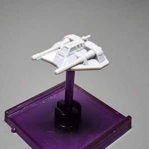 May include: A small, light gray model of a spacecraft, mounted on a purple square base with a black stem. The spacecraft has a detailed design with wings and a cockpit. The base is translucent, and the overall aesthetic is futuristic.