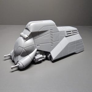 May include: A gray, futuristic-looking model of a vehicle with a rounded front and angular body. The model has detailed paneling, vents, and two protruding gun-like structures. It is set against a gray background.