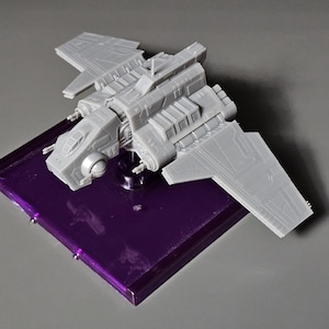 Könnte beinhalten: Ein graues Plastikmodell eines Raumschiffs mit Flügeln, das auf einem violetten Präsentationsständer steht.