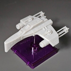 May include: A grey plastic model of a spaceship on a purple display stand. The spaceship has two engines and a long, curved body.