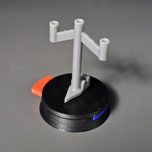 Könnte beinhalten: Ein grauer Kunststoff-Präsentationsständer mit drei Armen und einem schwarzen Sockel. Der Ständer ist für die Präsentation von kleinen Objekten wie Figuren oder Actionfiguren konzipiert.