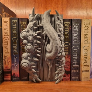 Könnte beinhalten: Ein grauer, 3D-gedruckter Buchstütze in Form eines Monsters mit mehreren Augen und Zähnen. Der Buchstütze hält einen Stapel Bücher mit dem Autor Bernard Cornwell auf den Rückseiten.