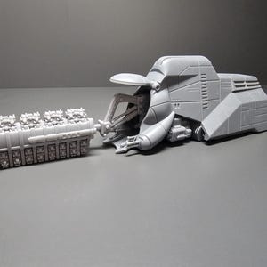 May include: A gray, futuristic-looking model of a vehicle with a large engine component. The vehicle has a streamlined body with detailed panels and a complex mechanical arm. The engine is a rectangular block with multiple cylinders.