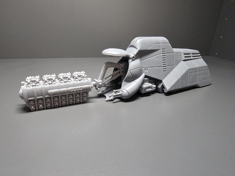 May include: A gray, futuristic-looking model of a vehicle with a large engine component. The vehicle has a streamlined body with detailed panels and a complex mechanical arm. The engine is a rectangular block with multiple cylinders.
