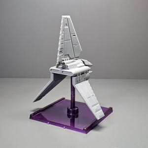 May include: A grey model spacecraft with angled wings and a vertical fin, mounted on a purple rectangular base. The spacecraft has detailed paneling and a central body. The base is a translucent purple colour.