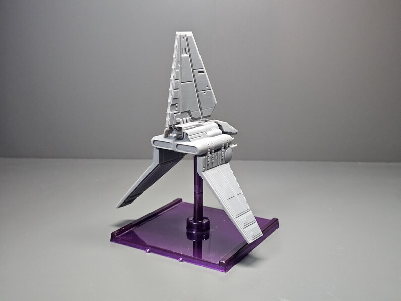 May include: A grey model spacecraft with angled wings and a vertical fin, mounted on a purple rectangular base. The spacecraft has detailed paneling and a central body. The base is a translucent purple colour.