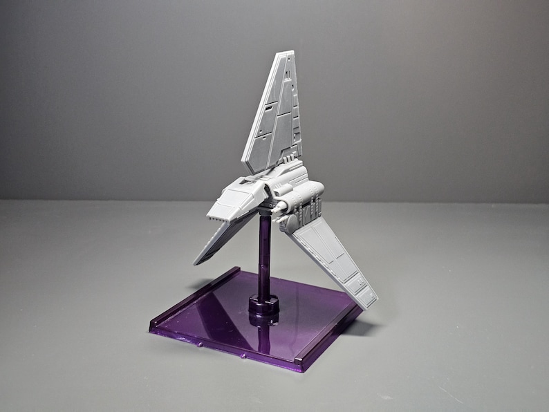 May include: A grey model spacecraft with angled wings and a vertical stabiliser, mounted on a purple rectangular base. The model is supported by a dark purple stand. The background is a solid grey.