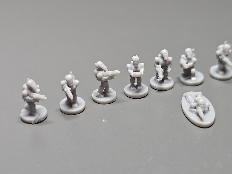 K&ouml;nnte beinhalten: Eine Sammlung kleiner, grauer Miniaturfiguren, die futuristische Soldaten darstellen. Die Figuren stehen auf kreisf&ouml;rmigen Sockeln, einige halten Waffen. Die Miniaturen sind auf einer grauen Oberfl&auml;che angeordnet.
