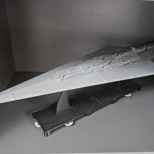 May include: A grey model of a Star Destroyer spaceship, displayed on a black stand. The model has detailed surface textures and is inside a grey cabinet. The spaceship's design features a long, triangular shape with intricate details.