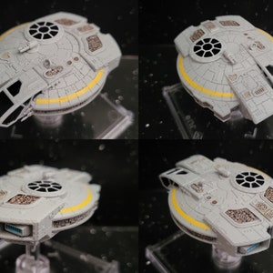 May include: A grey and white spaceship model with yellow accents. The model is on a clear display stand. The spaceship has a round body with a flat top and a pointed bottom. The model is detailed with various panels and hatches.