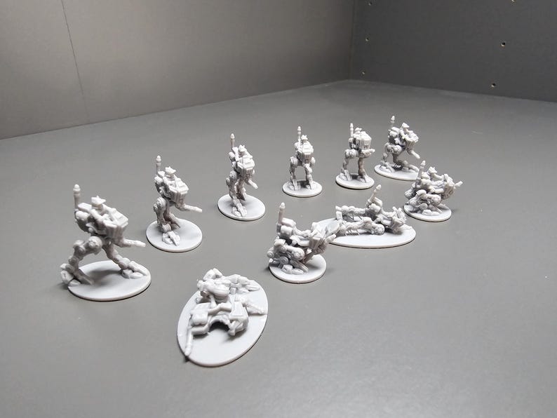 May include: A collection of small, gray, 3D-printed miniatures, likely for tabletop gaming. The figures include various robotic or futuristic designs, some standing upright and others in a more terrain-like form. Each miniature is on a small, round or oval base.