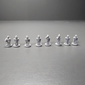 May include: Eight small, grey robot figurines are arranged in a row. Each robot stands on a small, circular base. The robots have detailed designs, with visible armour-like plating and helmets. The figurines are evenly spaced and appear to be plastic.