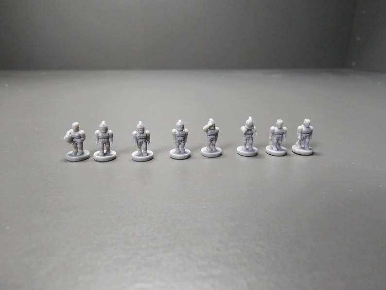 May include: Eight small, grey robot figurines are arranged in a row. Each robot stands on a small, circular base. The robots have detailed designs, with visible armour-like plating and helmets. The figurines are evenly spaced and appear to be plastic.