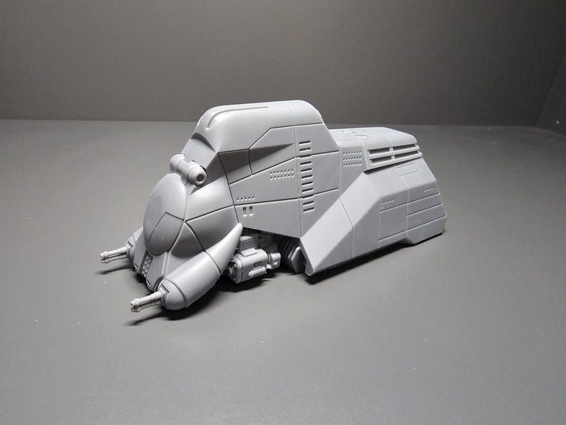 May include: A gray, futuristic-looking model of a vehicle with a rounded front and angular body. The model has detailed paneling, vents, and two protruding gun-like structures. It is set against a gray background.