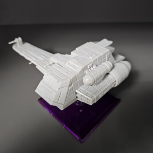 May include: A white plastic model of a spaceship with a purple base. The spaceship has a long, narrow body with a large, rectangular cockpit and a smaller, rounded engine section.