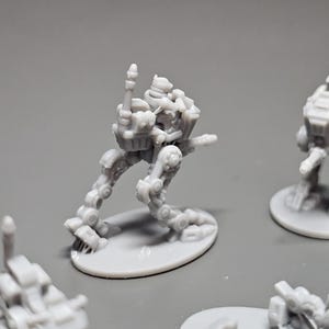 May include: Several small, gray, miniature robot figures are arranged on a gray surface. The robots have articulated limbs and detailed mechanical designs. One robot is in a dynamic pose, with its arm raised. The figures are on oval bases.