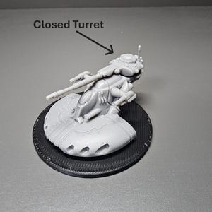 May include: A gray, miniature model of a closed turret vehicle, mounted on a black circular base. The model has detailed weaponry and a futuristic design. The text "Closed Turret" is above the model.