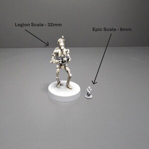 K&ouml;nnte beinhalten: Zwei Miniaturfiguren, eine 32 mm hohe und eine 8 mm hohe, werden auf einer grauen Oberfl&auml;che gezeigt. Die gr&ouml;&szlig;ere Figur ist ein detaillierter roboterartiger Charakter, w&auml;hrend die kleinere eine vereinfachte Version ist. Text zeigt die Skalierung.