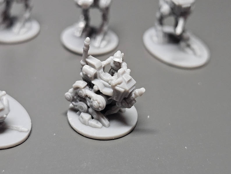 May include: A gray miniature figurine of a futuristic vehicle with a figure in the driver's seat. The vehicle is mounted on a circular base, with other similar figurines in the background. The details of the vehicle are intricate.