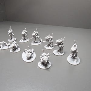 May include: A collection of small, gray, miniature robot figures. The figures are in various poses, some standing, some crouching, and some lying down. Each figure is on a small, round base. The background is a plain, light gray surface.