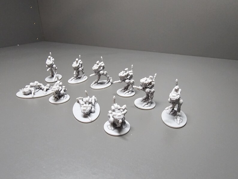 May include: A collection of small, gray, miniature robot figures. The figures are in various poses, some standing, some crouching, and some lying down. Each figure is on a small, round base. The background is a plain, light gray surface.