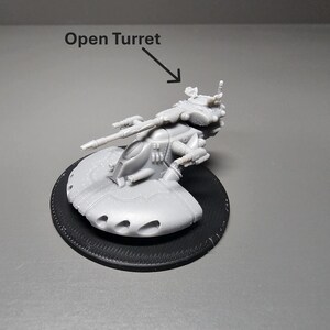 May include: A gray plastic model of a futuristic vehicle with an open turret, mounted on a black circular base. The model has detailed features, including a long gun barrel and multiple small holes. The text "Open Turret" is above the model.