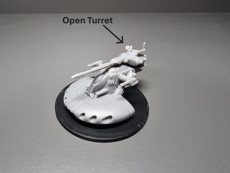 May include: A gray plastic model of a futuristic vehicle with an open turret, mounted on a black circular base. The model has detailed features, including a long gun barrel and multiple small holes. The text "Open Turret" is above the model.