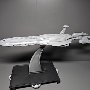 May include: A grey spaceship model, displayed on a black stand with a curved support. The spaceship features a long, sleek body with detailed elements, including wings and several smaller attached craft. The stand has a rectangular base.