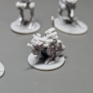May include: A collection of small, gray miniature figurines, likely for tabletop gaming or model building. The central figure is a detailed, mechanical-looking vehicle on a circular base, with other figures in the background.