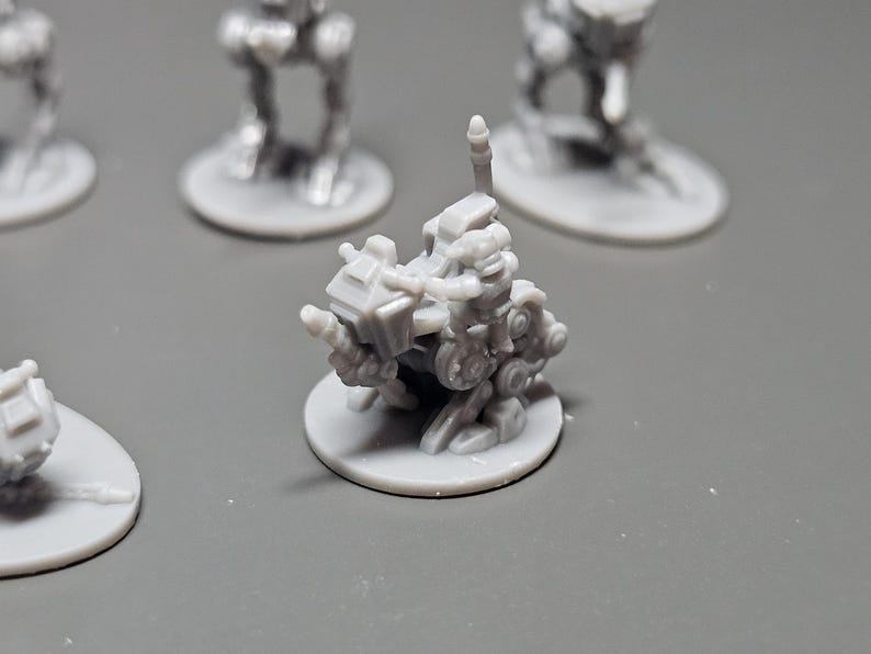 May include: A collection of small, gray miniature figurines, likely for tabletop gaming or model building. The central figure is a detailed, mechanical-looking vehicle on a circular base, with other figures in the background.