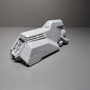 May include: A gray, futuristic-looking model of a vehicle with a cockpit, angled panels, and detailed mechanical elements. The model has a textured surface and is set against a gray background.