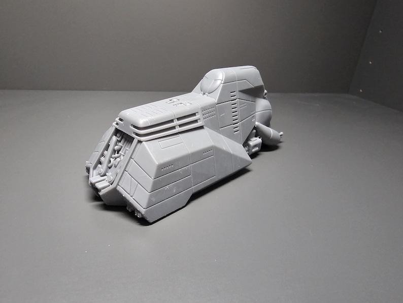 May include: A gray, futuristic-looking model of a vehicle with a cockpit, angled panels, and detailed mechanical elements. The model has a textured surface and is set against a gray background.