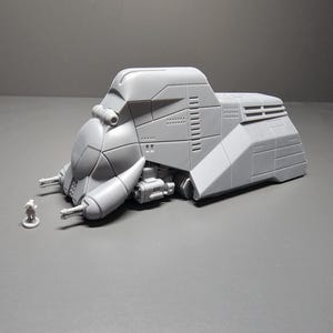 May include: A gray, futuristic vehicle model with a detailed design, featuring a rounded front, side-mounted protrusions, and a small figure for scale. The model has a textured surface and is set against a gray backdrop.